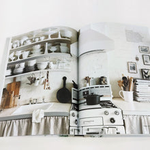 Load image into Gallery viewer, Curate book opened with pictures of white shelves with plates, bowls, and dishware above a counter and stove with black pan.